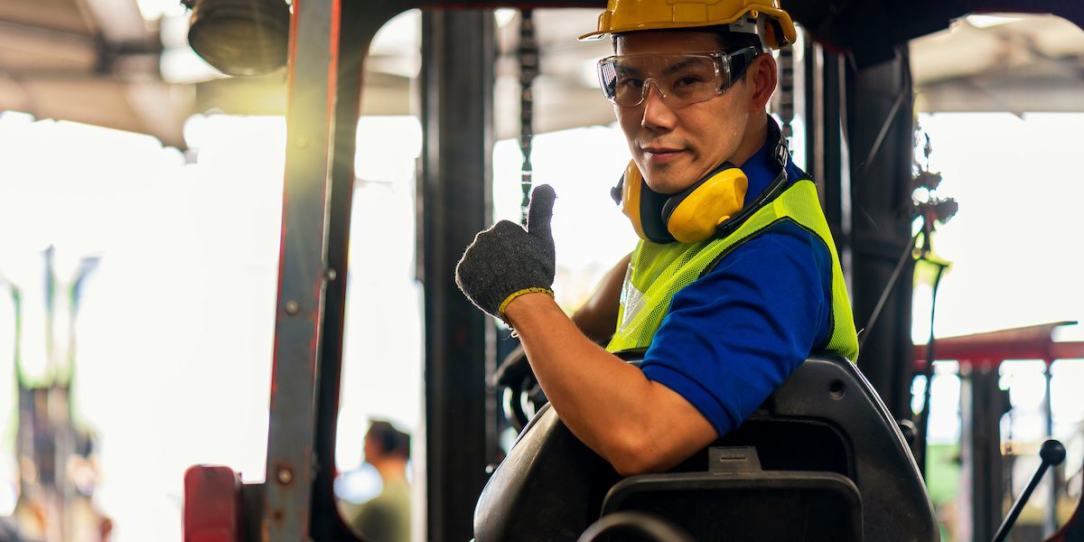 Engineer or technician Concept. A male employee driving a forklift and showing thumb up in factory.